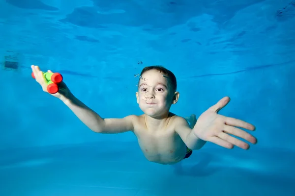 Retrato de un chico que se sumerge alegremente en el fondo de la piscina para juguetes. Sonríe y mira dentro del marco de la cámara. Buceos divertidos bajo el agua. Clase de natación. Niño feliz activo. Estilo de vida saludable. — Foto de Stock