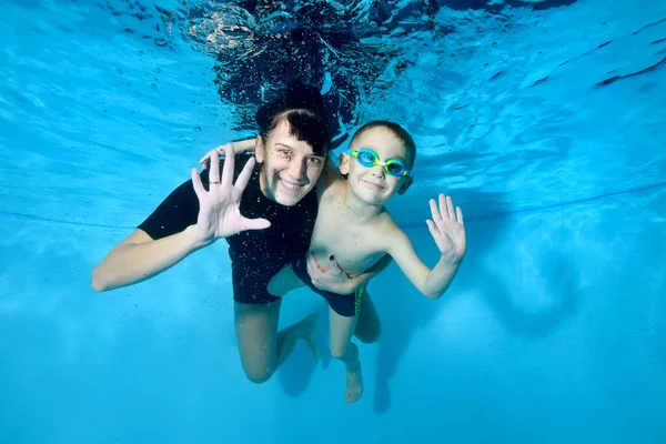 Retrato de un hermoso niño y su entrenador femenino bajo el agua. Sonríen, miran a la cámara y saludan. Niño feliz activo. Clases de natación para niños pequeños. Estilo de vida saludable — Foto de Stock