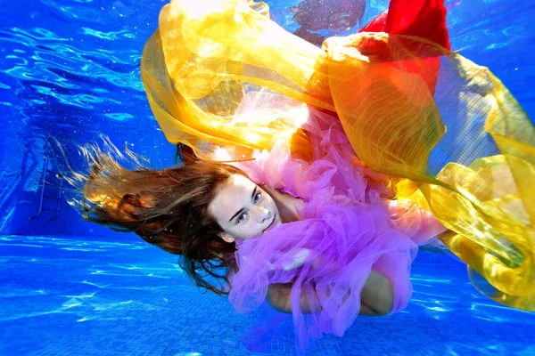 Portret van een charmant meisje met lang haar dat onder water zwemt en speelt in een zwembad met gele en rode stoffen. Modeportret. Onderwaterfotografie. Horizontale oriëntatie. — Stockfoto