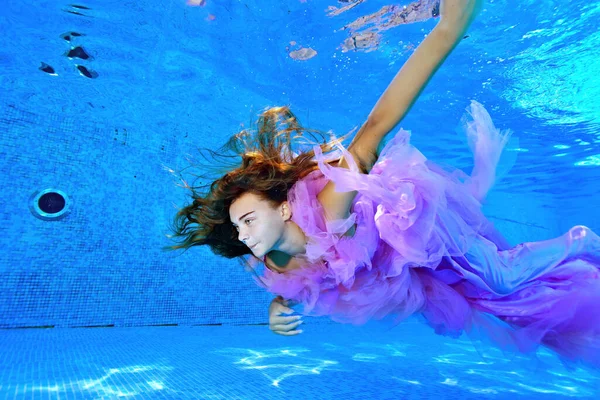 Una joven con un hermoso vestido nada bajo el agua en una piscina al aire libre con los brazos extendidos. Su pelo largo revolotea en el agua. Retrato. Ángulo ancho. Orientación horizontal. — Foto de Stock