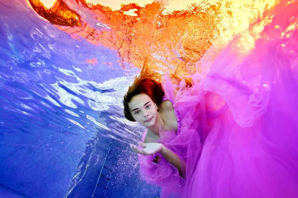 Een schattig meisje in een weelderige roze jurk duikt onder water in een buitenzwembad. Ze kijkt naar de camera tegen de achtergrond van de zonsondergang op het oppervlak. Onderwaterfotografie. Uitzicht vanaf de onderkant. — Stockfoto