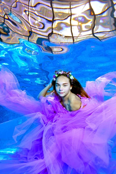 Egy szürreális portré egy tinédzser lányról. Belenéz a kamerába, és a víz alatt pózol egy kültéri medence alján, lila ruhában és koszorúban. Víz alatti divat fotózás. — Stock Fotó