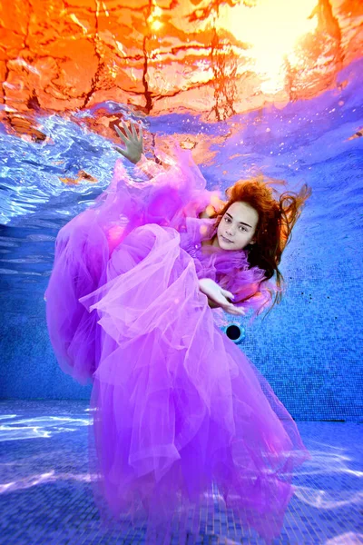 Onderwaterportret van een jong meisje in een paarse jurk die poseert voor de camera onder water in een zwembad tegen een achtergrond van geel licht. Heldere kleuren. Portret. Modieuze onderwaterfotografie. — Stockfoto