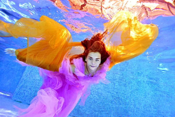 En vacker ung flicka simmar under vattnet i en pool i en lila klänning och med en gul trasa i händerna. Hennes armar är utspridda som vingar. Undervattens mode fotografering. — Stockfoto