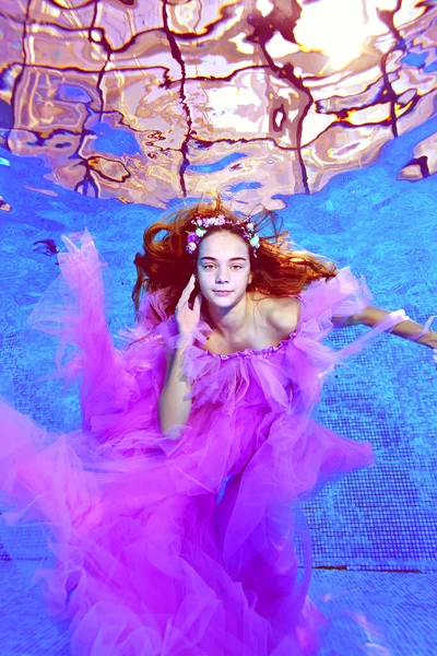 Un portrait branché d'une belle fille en robe violette posant et regardant la caméra sous l'eau au fond d'une piscine extérieure. Photographie sous-marine. Orientation verticale. — Photo