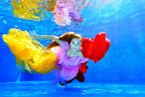 Een jong meisje met lang haar zwemt en poseert onder water in een buitenbad met kleurrijke stoffen in haar handen. Modeportret. Onderwaterfotografie. Horizontale oriëntatie. — Stockfoto