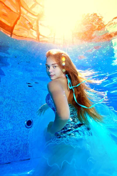 Um retrato subaquático incomum de uma bela jovem que posa em um belo vestido subaquático em uma piscina exterior em um dia ensolarado brilhante. Ela olha por cima do ombro para a câmara.. — Fotografia de Stock