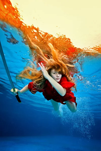 Una hermosa joven con el pelo suelto nada bajo el agua en un vestido rojo y con una espada samurai en la mano y mira a la cámara. Fotografía bajo el agua. Retrato de moda. —  Fotos de Stock