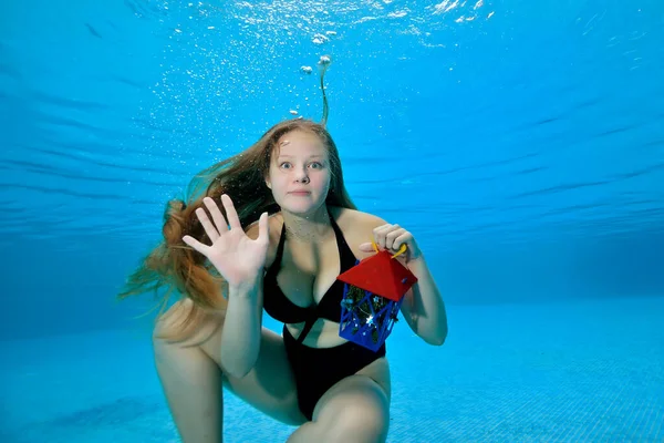 Sexy chica posando para la cámara bajo el agua en la piscina con un regalo en la mano en un día soleado sobre un fondo azul. Retrato. El concepto. Disparo creativo. Ángulo ancho. —  Fotos de Stock