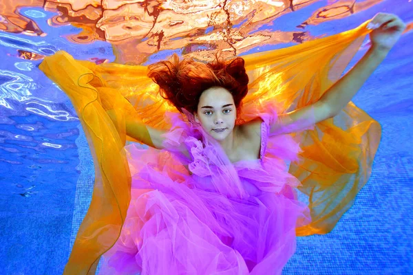 Uma adolescente encantadora posa debaixo d 'água em uma piscina com um pano amarelo em suas mãos em um vestido roxo em um fundo azul e olha para a câmera. Um retrato conceptual. Vista horizontal. — Fotografia de Stock