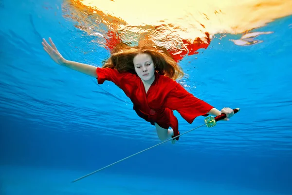 Ένα μοδάτο πορτραίτο ενός κοριτσιού με κόκκινο κιμονό, που κολυμπάει και ποζάρει κάτω από το νερό σε μια πισίνα με ένα σπαθί σαμουράι στο χέρι της σε μια ηλιόλουστη μέρα σε μπλε φόντο. Δημιουργικός πυροβολισμός. Έννοια. Ευρεία γωνία. — Φωτογραφία Αρχείου