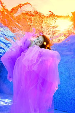 Suyun yüzeyinden gelen gerçeküstü ışığa karşı, gözleri suyun altında, mor bir elbiseyle uyuyan genç bir kız. Su altı konsepti fotoğrafçılığı. Moda portresi.