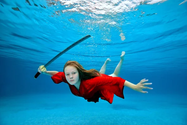 Una chica agresiva en un kimono rojo simula un ataque con una espada samurai mientras nada bajo el agua en una piscina al aire libre en aguas cristalinas. Disparo creativo. Concepto. Retrato de una mujer. Ángulo ancho. — Foto de Stock