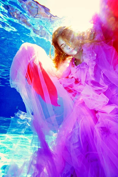 Ett surrealistiskt porträtt av en tonåring flicka under vatten i en lyxig flerfärgad klänning mot bakgrund av starkt solljus från ytan av vattnet. Undervattensfotografering. — Stockfoto