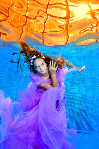 Una joven adolescente se divierte y nada en la piscina con un hermoso vestido morado con el pelo suelto y una corona de flores en la cabeza, y mira a la cámara. Retrato de moda. Concepto. —  Fotos de Stock