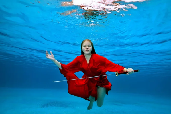 Una hermosa chica en un kimono rojo posa para la cámara bajo el agua en una piscina con una espada samurai en un día soleado sobre un fondo azul. Retrato de moda. Disparo creativo. Ángulo ancho. Copiar espacio. —  Fotos de Stock