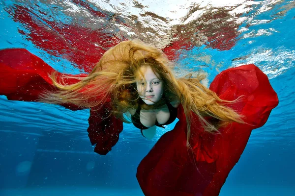 Um retrato incomum de uma jovem com cabelo luxuoso sob a água na piscina. Ela nada e posa para a câmera com um pano vermelho, braços estendidos. Seu cabelo flutua na água. — Fotografia de Stock