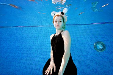 Çekici bir kız, güneşli bir günde siyah bir elbiseyle havuzun dibinde oturuyor. Kafasında beyaz boynuzları var ve mavi arka planda kameraya poz veriyor. Konsept. Geniş açı.