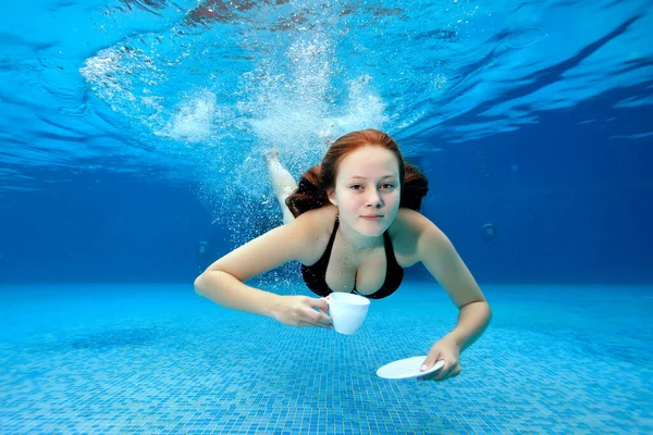 Sexy chica nada y posa bajo el agua en la piscina y sostiene una taza blanca y platillo en sus manos. Ella nada en un traje de baño negro en un día soleado y mira a la cámara. Retrato de moda. Ángulo ancho. —  Fotos de Stock