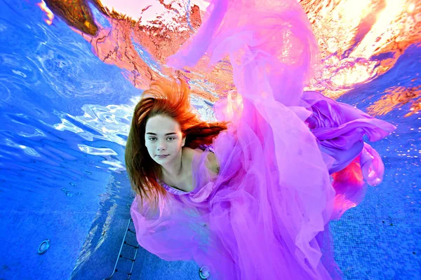 Retrato de uma adolescente encantadora debaixo d 'água em um vestido roxo luxuoso contra a luz surreal da superfície da água. Fotografia de conceito subaquático. Retrato de moda. Vista vertical. — Fotografia de Stock