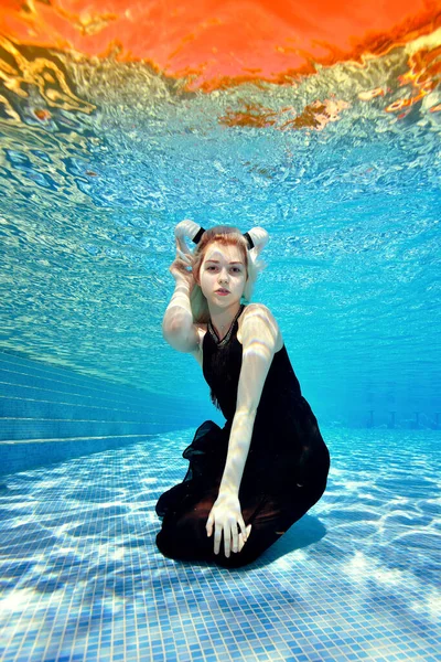 在阳光灿烂的一天，一个金发碧眼的女孩身穿黑色连衣裙，头戴白角，坐在室外游泳池底部，在水下对着摄像机摆姿势。时尚摄影. — 图库照片