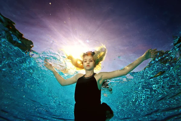 Un retrato surrealista de una joven rubia nadando bajo el agua en una piscina con un vestido negro, brazos extendidos como un pájaro. Disparo desde el fondo de la piscina en la luz del contorno del sol. —  Fotos de Stock