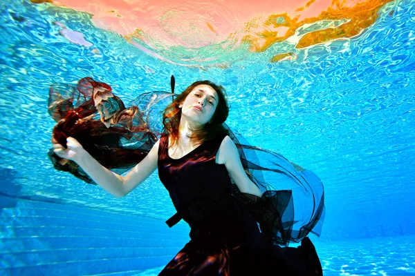 Een mooi meisje danst onder water met een rode doek in haar handen in een rode jurk op een heldere zonnige dag. Modieuze onderwaterfotografie. Het concept. Horizontale oriëntatie. — Stockfoto