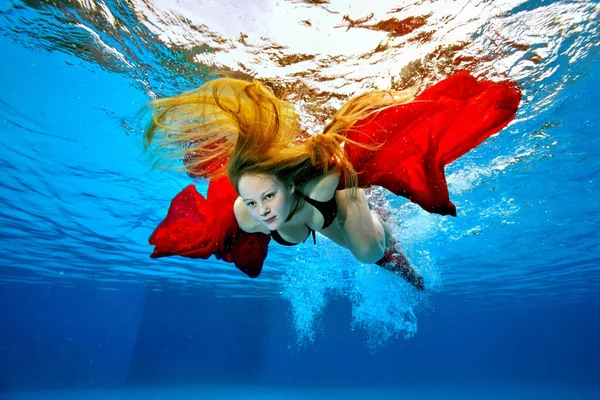 Een badmeester meisje met lang haar duikt onder water en zwemt met een rode doek in haar handen op een stralende zonnige dag. Portret. Onderwaterfotografie. Onderaanzicht. Horizontale oriëntatie. — Stockfoto