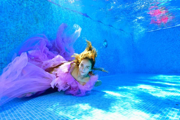 Uma jovem em um lindo vestido roxo fica debaixo da água no fundo da piscina com o cabelo para baixo e olha para a câmera. Retrato de moda. Fotografia subaquática criativa. Ângulo largo. — Fotografia de Stock