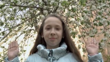 Bir bahar günü bahçede çiçek açan bir ağacın arka planında oynayan tatlı bir genç kız. Kapatıyor ve elleriyle gözlerini açıyor, kameraya bakıyor ve gülümsüyor. Portre.