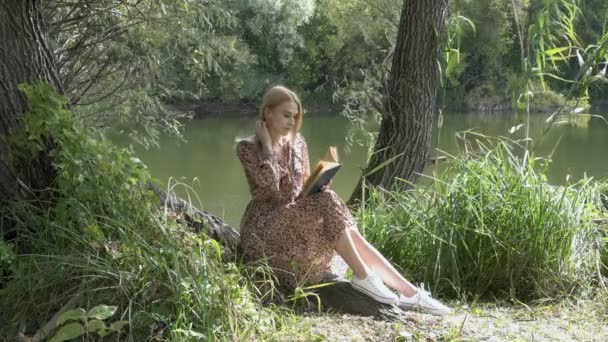 Een mooi studentmeisje zit op het gras in de natuur bij het water, leest een boek, raakt haar haar aan en kijkt rond. Concept. Modeportret. 4k. — Stockvideo
