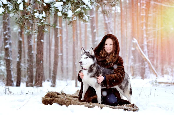 Retrato de una hermosa niña en un abrigo de piel en un día de invierno con un perro Husky en el parque. La chica abraza suavemente al perro y mira a la cámara. Sobre nieve blanca con un perro. Personas y perros. — Foto de Stock