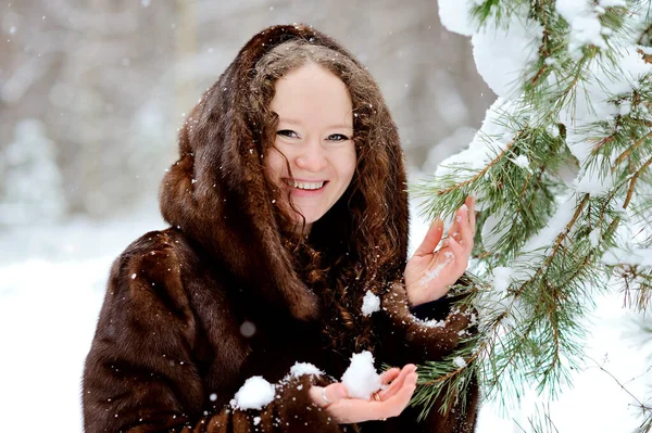 공원의 어느 화창 한 겨울날 모피 후드를 입은 아름다운 소녀의 모습. 그 소녀는 카메라 를 부드럽게 바라본다. 눈 속의 전나무의 가지들. — 스톡 사진