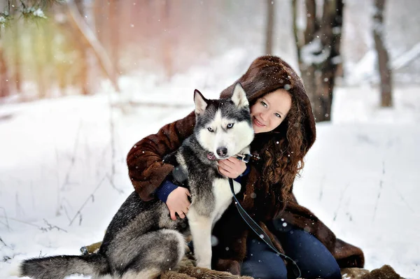 Retrato de una hermosa niña en un paseo en un día soleado de invierno con un perro Husky en el parque. La chica abraza suavemente al perro y mira a la cámara. Un hombre sobre nieve blanca con un perro. Personas y perros. — Foto de Stock