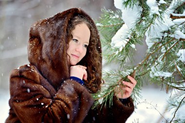 Parktaki güneşli bir kış gününde kürk giyen genç ve tatlı bir kızın görüntüsü. Kız köknar ağacının dallarına şefkatle bakıyor. Kardaki köknar ağaçlarının dalları. Yatay yönelim.