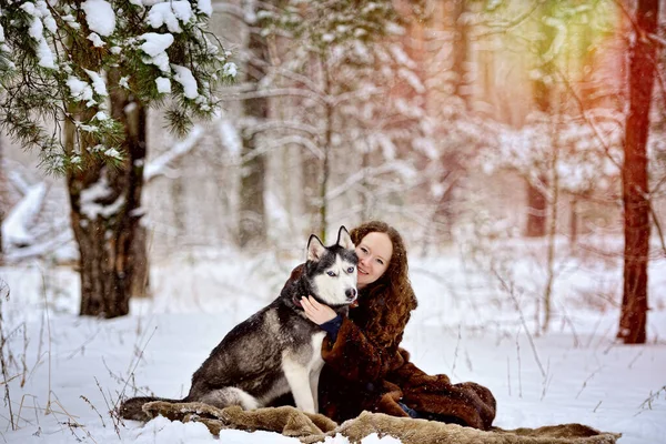 Imagen de una joven sonriente en un día soleado de invierno en el bosque con un perro Husky. Una mujer está sentada al lado de un perro. Una chica con un hermoso abrigo de piel. Un hombre sobre nieve blanca con un perro. Personas y perros. — Foto de Stock