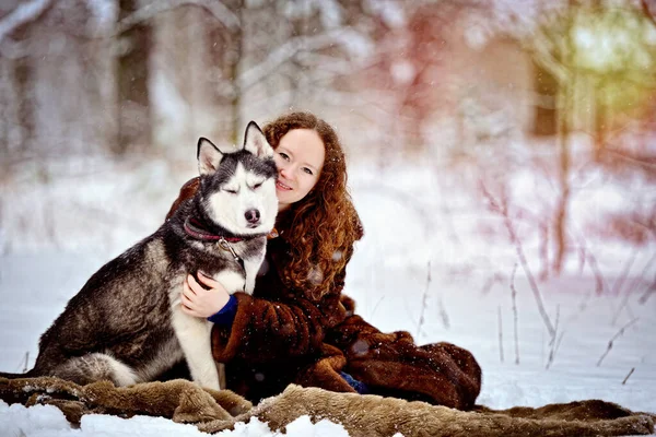 Imagen de una joven sonriente en un día soleado de invierno en el bosque con un perro Husky. Se acurrucó con el perro. Una chica con un hermoso abrigo de piel. Un hombre sobre nieve blanca con un perro. Personas y perros. — Foto de Stock