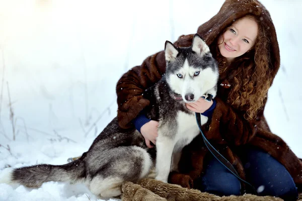 Retrato de una joven en una capucha de piel con un perro Husky en un parque de invierno. Un hombre sobre nieve blanca con un perro. Personas y perros. Orientación horizontal. — Foto de Stock