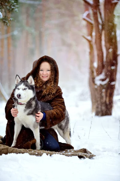 Retrato de una hermosa niña en un abrigo de piel en un paseo en un día soleado de invierno con un perro Husky en el parque. La chica abraza suavemente al perro y mira a la cámara. Sobre nieve blanca con un perro. — Foto de Stock