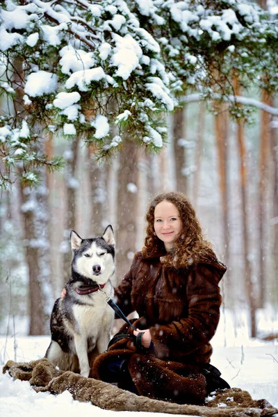 Retrato de una joven atractiva en un día soleado de invierno en el bosque con un perro Husky. Está sentada al lado del perro. Una chica con un hermoso abrigo de piel. Un hombre sobre nieve blanca con un perro. — Foto de Stock