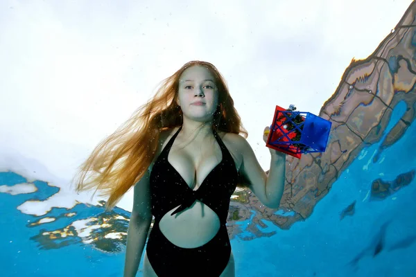 Retrato de una joven con el pelo largo nadando bajo el agua en una piscina con un juguete en la mano en un traje de baño negro en un día soleado sobre un fondo azul. El concepto. Ángulo ancho. Vista inferior. —  Fotos de Stock
