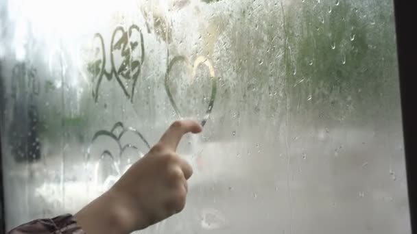 Дитина малює серце пальцем на затупленому склі автобуса, який подорожує містом у похмурий, дощовий день. Автомобілі їдуть за вікном. Поняття. Безликий. Крупним планом. 4-кілометровий — стокове відео