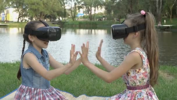 Kinderen, twee jonge meisjes, spelen in virtual reality glazen, zittend in een park tegen de achtergrond van water op een zomeravond. Ze houden elkaars handen vast en glimlachen. Het concept. Een close-up. 4K — Stockvideo