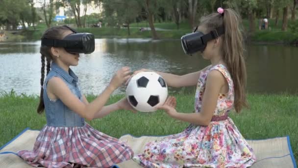 Dua gadis muda dalam kacamata virtual reality bersama-sama memutar dan mempelajari bola sepak dengan sentuhan, duduk di taman dengan latar belakang air pada hari musim panas. Konsep. Close-up. 4K — Stok Video