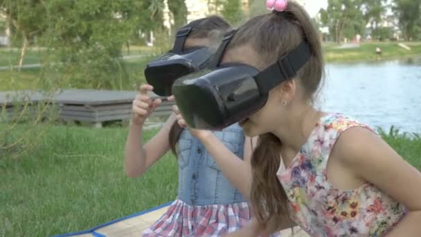 Close-up, twee jonge meisjes spelen virtuele games met een zwarte virtual reality bril, zittend in de achtertuin bij de rivier op een zomeravond. Het concept van een vakantie in quarantaine. 4K — Stockvideo