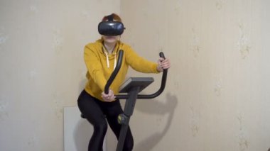 Sanal gerçeklik gözlüğü takan genç bir kız evde egzersiz bisikleti ile spor yapıyor ve kulaklıkla müzik dinliyor. Sarı spor kıyafetlerle pedal çeviriyor. Kavram. 4K