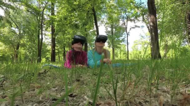 Маленькая дочь и её мать играют в виртуальные игры, лежа в очках виртуальной реальности на траве в парке летним вечером. Концепция. Стрельба с нижней точки. Широкий угол. 4K — стоковое видео