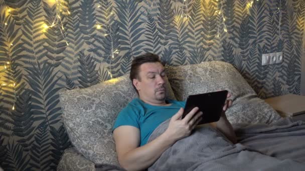 Un hombre de mediana edad descontento lee las noticias en una tableta mientras está acostado en la cama de su dormitorio por la noche, con una guirnalda parpadeante en el fondo. El concepto. Primer plano. 4K — Vídeo de stock