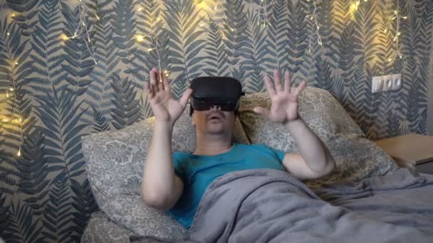 Un hombre juega juegos virtuales mientras está acostado en gafas de realidad virtual por la noche en una cama debajo de una manta sobre un fondo de guirnalda amarilla intermitente. Primer plano. Retrato. 4K — Vídeos de Stock