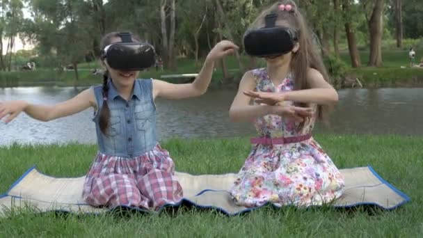 İki komik küçük kız, bir yaz akşamı parkta çimlerin üzerinde otururken sanal gerçeklik gözlükleriyle oynuyorlar. Ellerini sallıyorlar, hayali bir nehirde yüzüyorlar ve gülüyorlar. Portre. 4K — Stok video